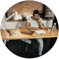 Man sitting at a desk working on a laptop and notebook. Phone up to his ear. 