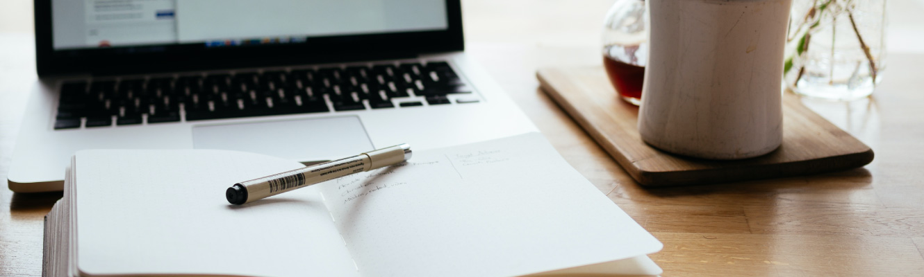 Close up of various items on a wooden surface, including a laptop and notebook with a pen